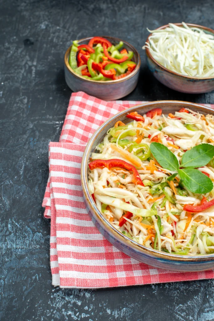 A colorful vegetable salad featuring shredded cabbage, red and green bell peppers, and carrots, served in a bowl on a red checkered napkin