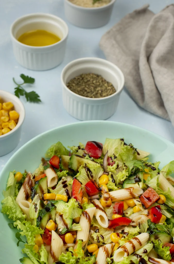 A close-up of a pasta salad with penne, corn, cucumbers, and balsamic drizzle, displayed alongside ingredients like olive oil and herbs. Not related to an elote salad.